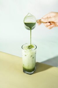 A refreshing iced matcha latte being poured from a small jug into a tall glass on a light surface.